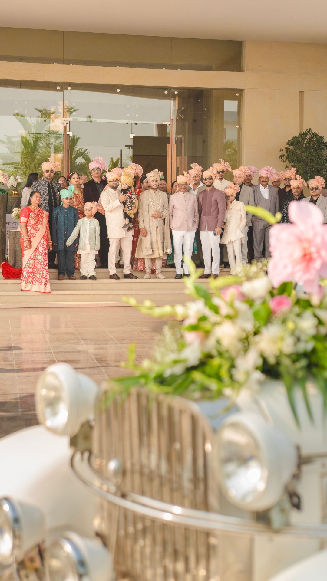 Indian wedding group photo with men in colorful turbans and sherwanis posed outside a luxury venue beside a vintage white car.