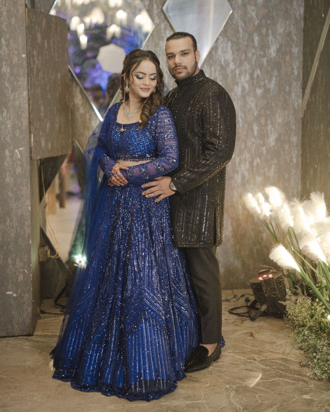 Couple in elegant Indian attire, woman in blue sequined lehenga, man in black sherwani, posing at a formal event.