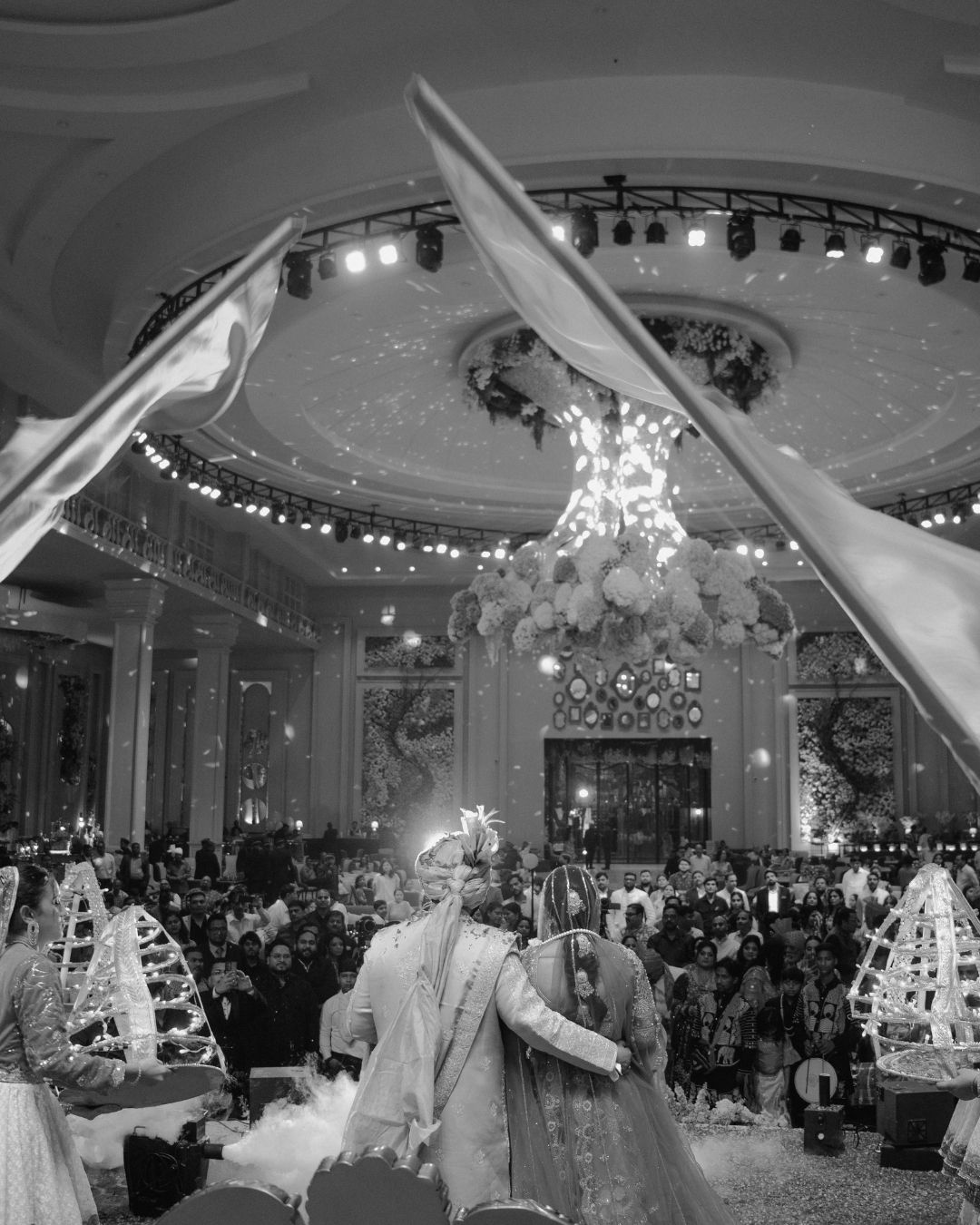 Black and white photo of an Indian wedding couple entering a grand ballroom with chandelier and guests.