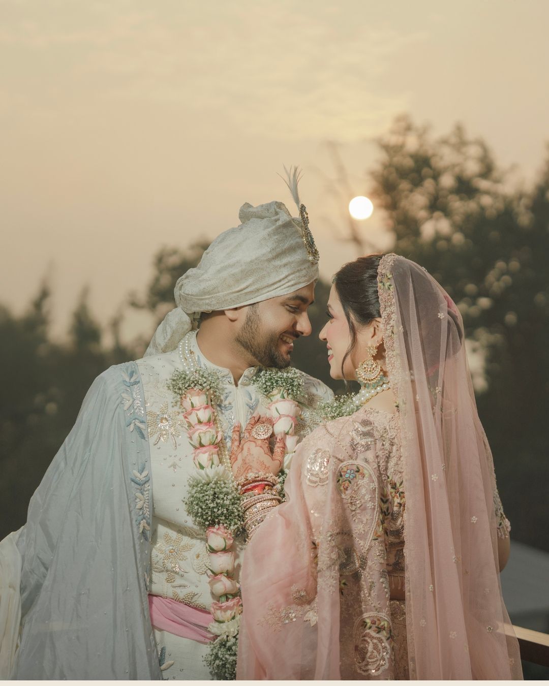 Indian bride and groom in traditional wedding attire exchange loving glances at sunset, adorned with floral garlands.