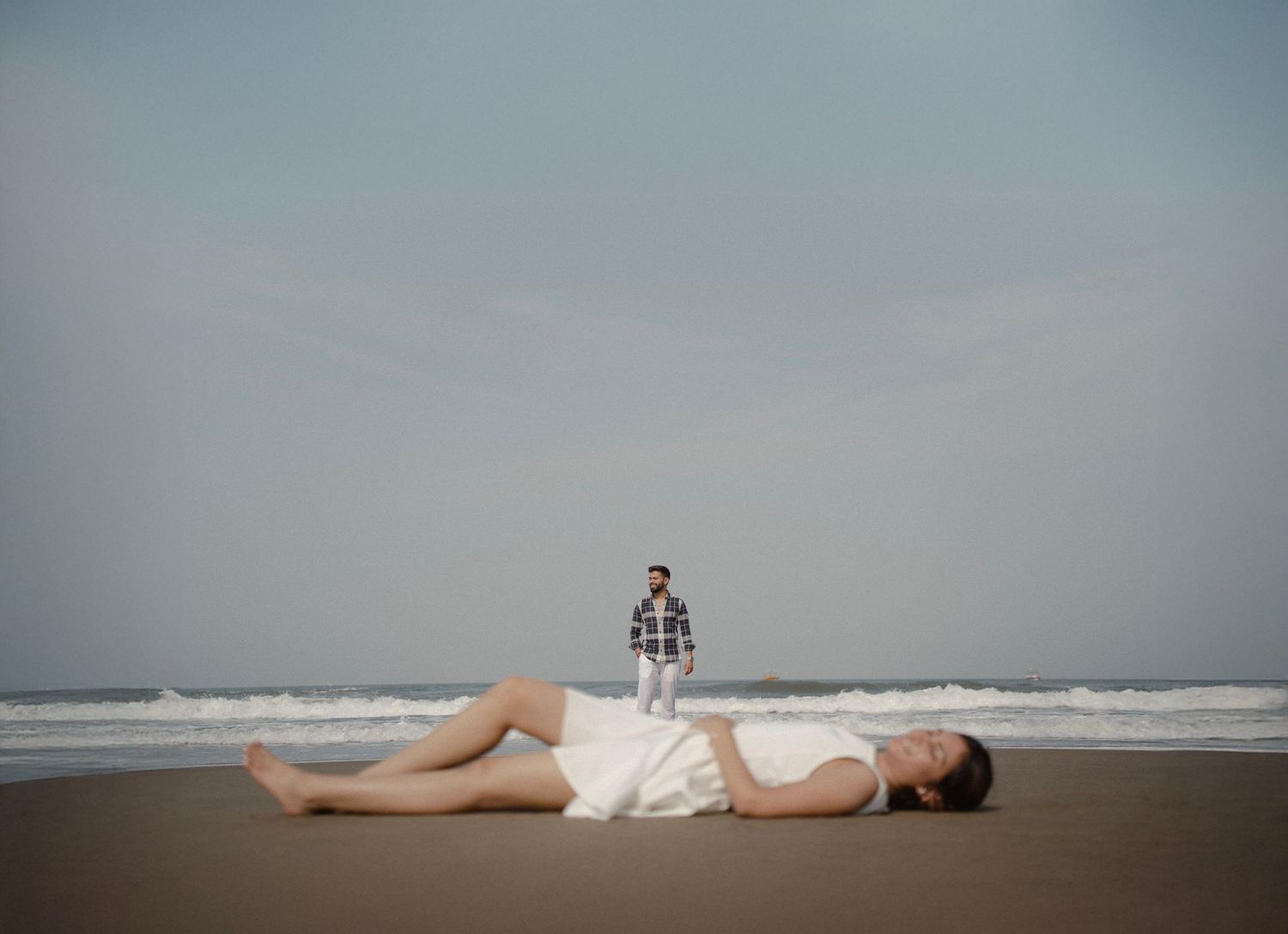 Woman lying on beach in white dress with man standing in background near ocean waves on overcast day.