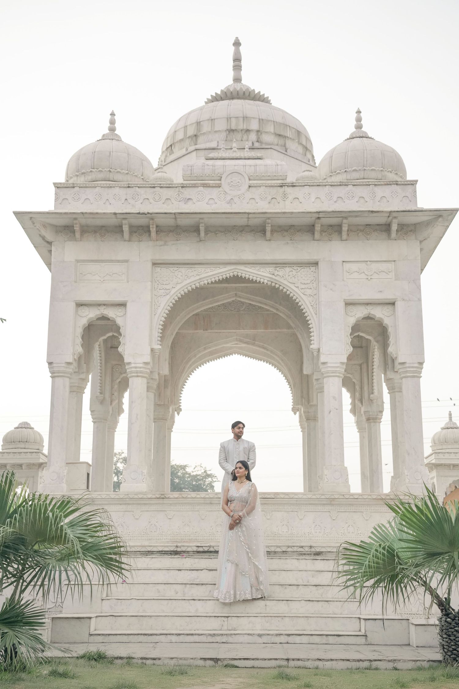 Couple in white attire posing on steps of ornate white marble Mughal-style pavilion with domed arches.