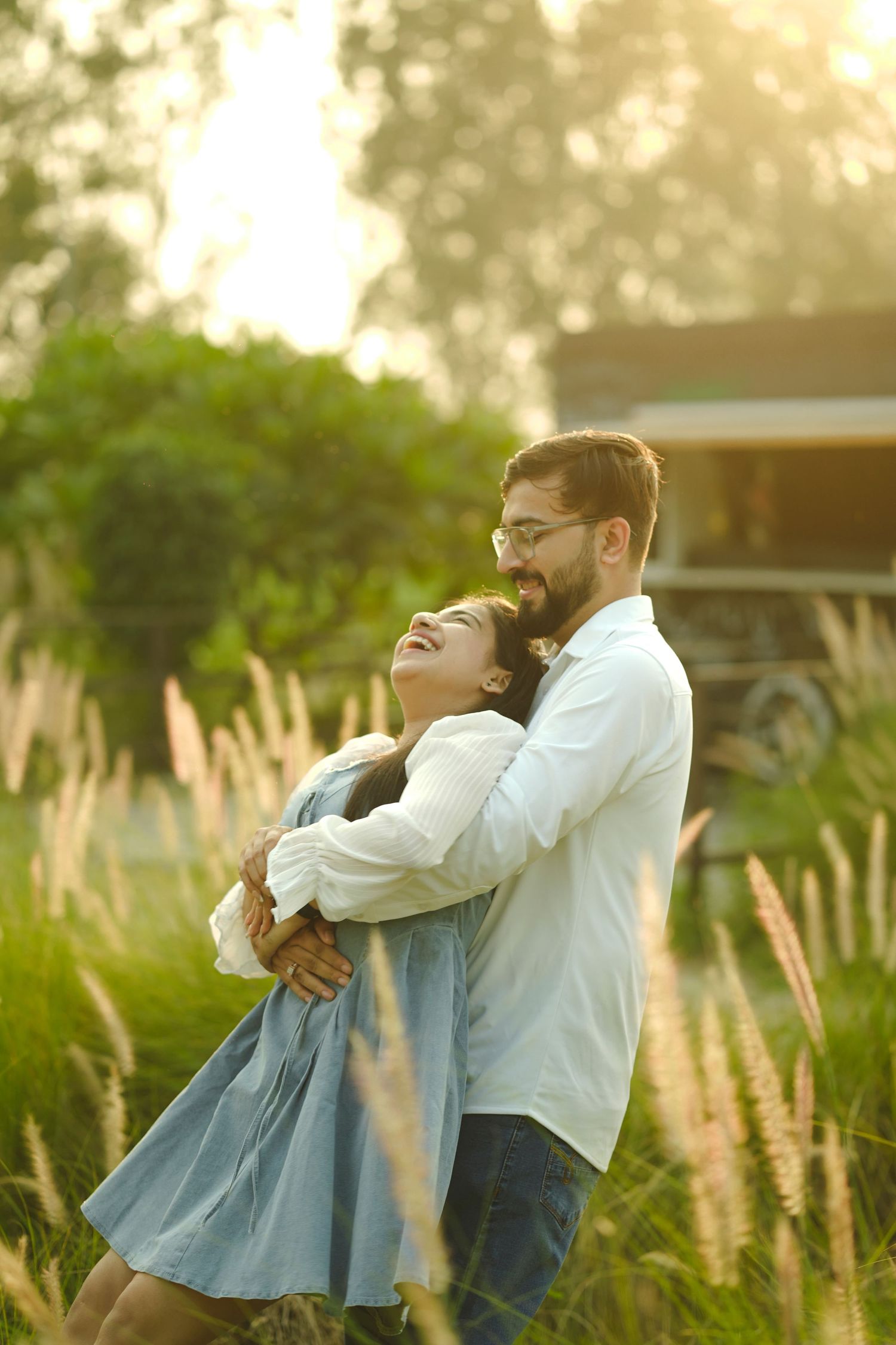 A couple embracing and laughing joyfully in a sunlit meadow with tall grass and green trees in the background.