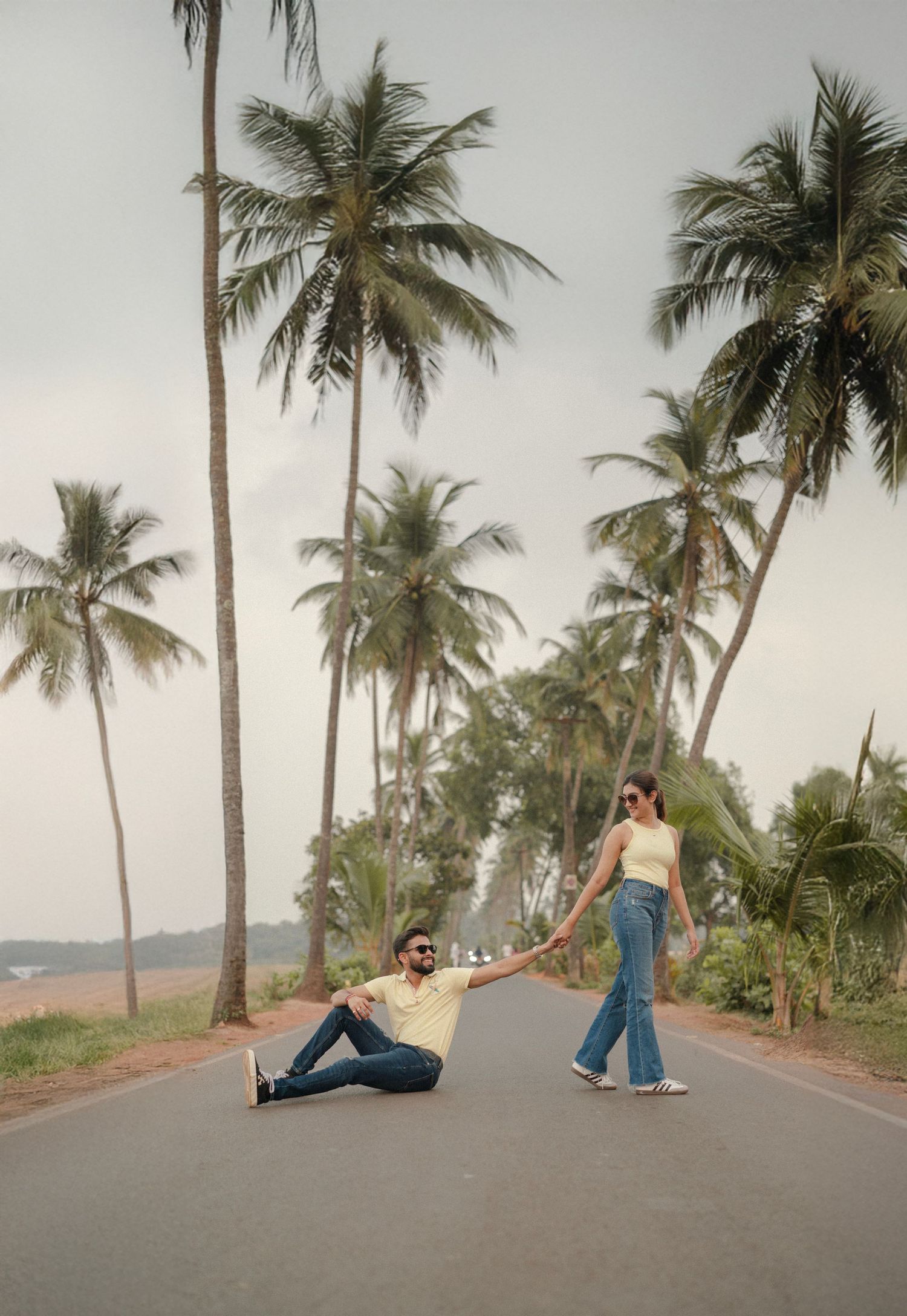 Couple holding hands on a palm-lined road, man sitting on ground, woman standing in jeans, tropical beach setting.