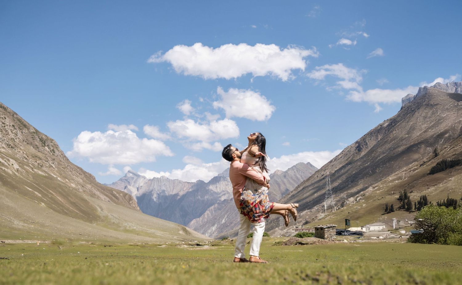Couple embracing in a scenic mountain valley under blue skies, surrounded by green meadows and snow-capped peaks.