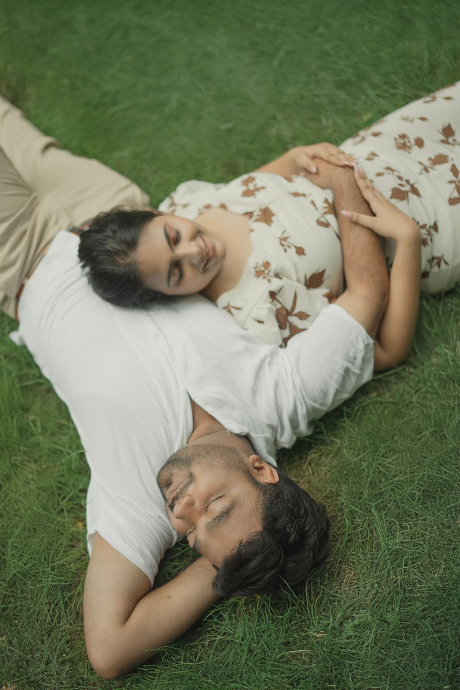 Couple lying on green grass, woman resting head on man's chest, sharing a peaceful romantic moment outdoors.