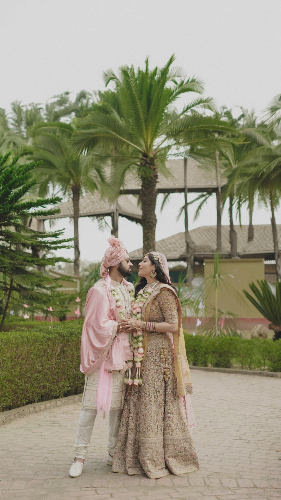 Indian bride and groom in traditional wedding attire standing together outdoors with palm trees in background.