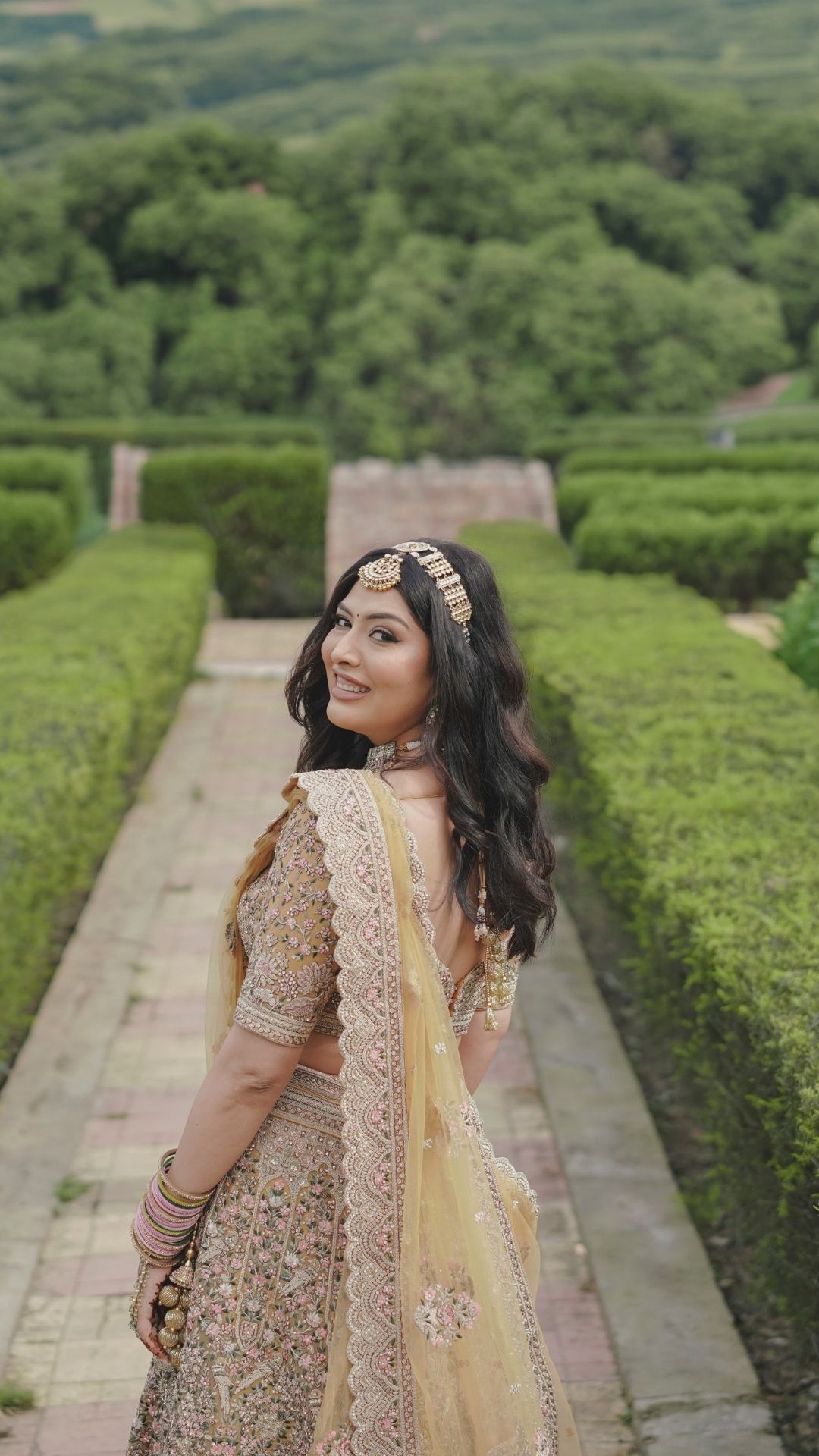 Smiling bride in golden lehenga with maang tikka poses in lush green garden pathway.