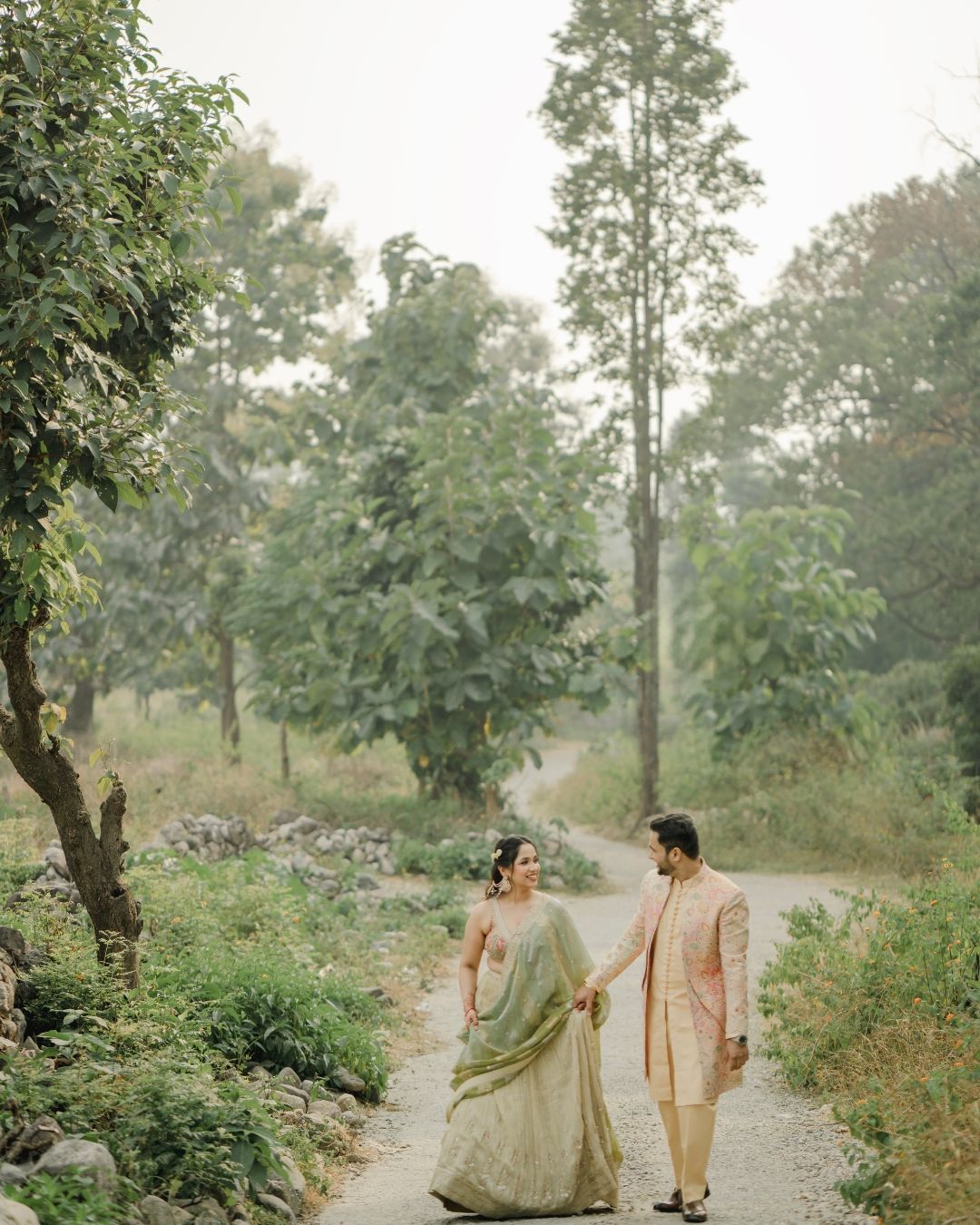 Indian couple in traditional wedding attire holding hands walking on a scenic forest path.