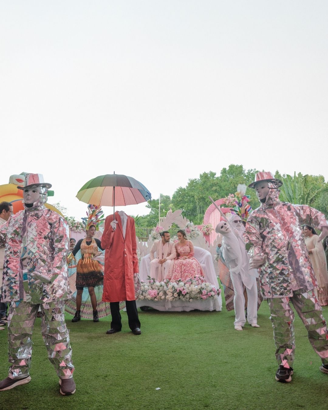 Colorful stilt walkers perform at an outdoor Indian wedding ceremony with couple seated under floral arch.