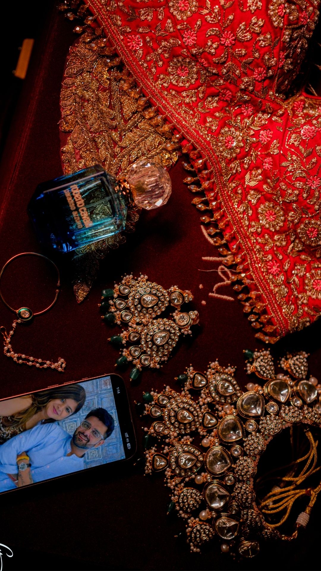 Bridal flat lay with red embroidered lehenga, kundan jewelry set, perfume bottle, and phone displaying couple photo.