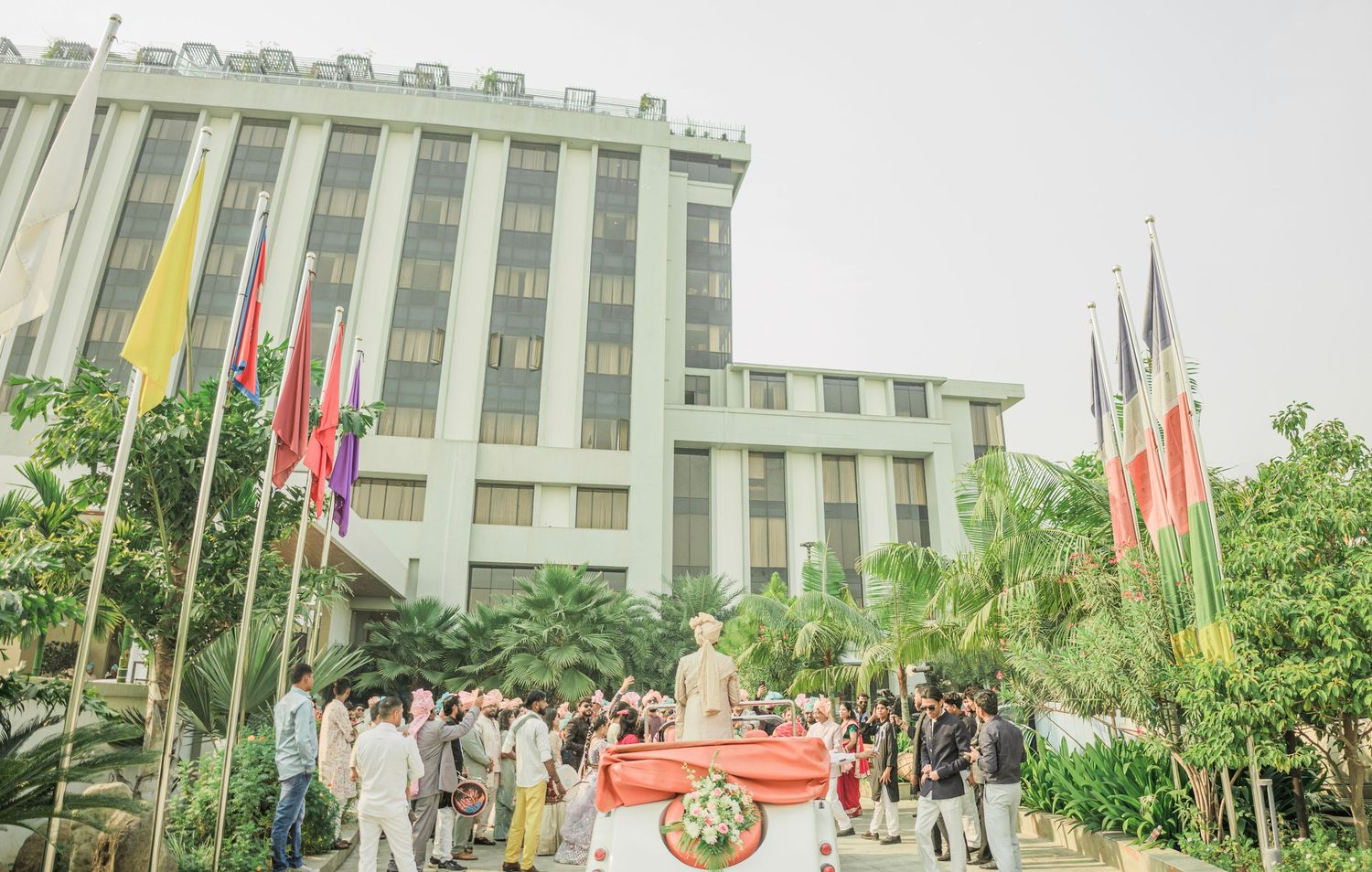 Crowd gathers around a statue in front of a large building with international flags, surrounded by tropical trees.