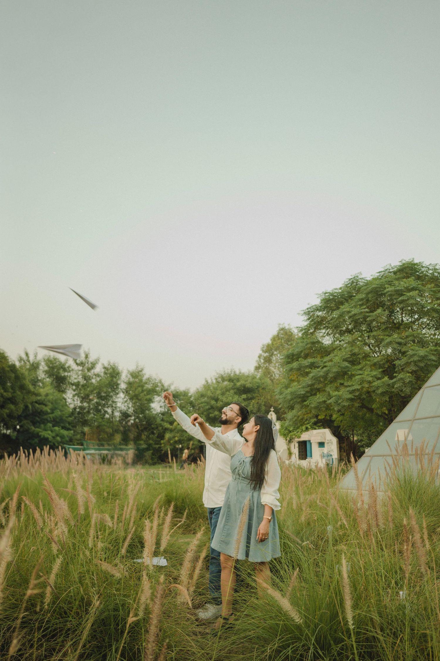 Couple standing in a meadow of tall grass, joyfully tossing a paper airplane into the sky near trees.