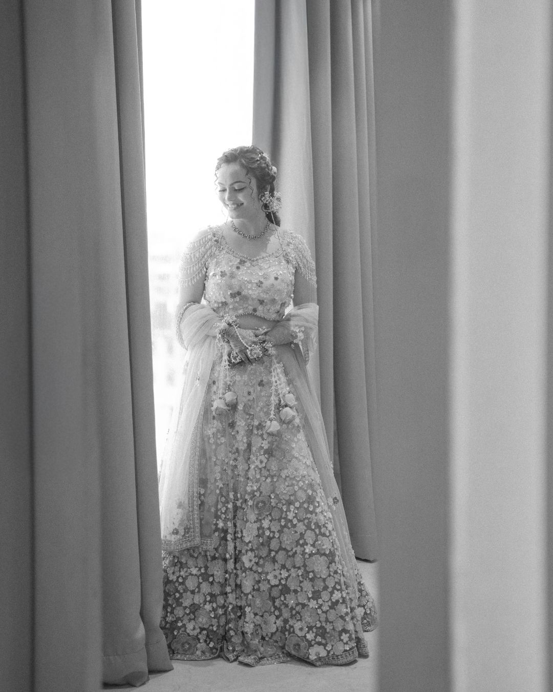Black and white photo of a bride in floral lehenga standing by curtained window on her wedding day.