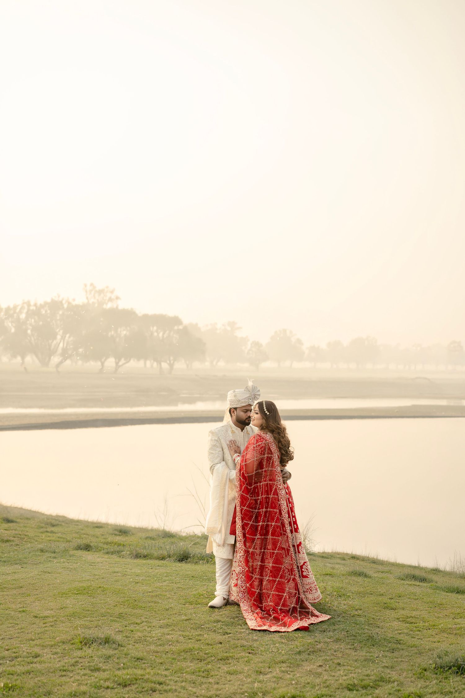 Indian bride in red saree and groom in white sherwani share a romantic moment by a misty lake.