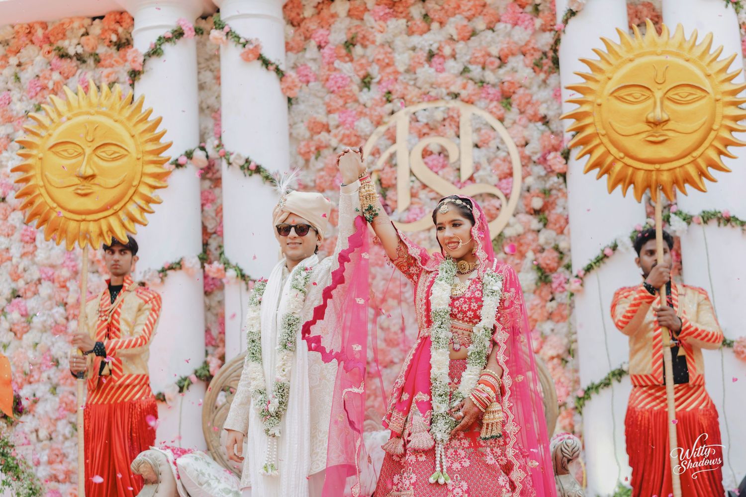 Traditional Indian wedding ceremony with decorative golden sun motifs and pink floral arrangements against white backdrop.