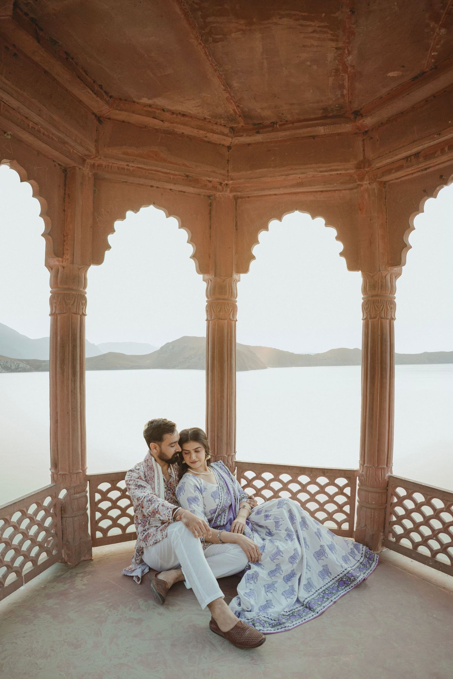 Couple in traditional Indian attire sitting in an ornate Rajasthani pavilion overlooking a serene lake.