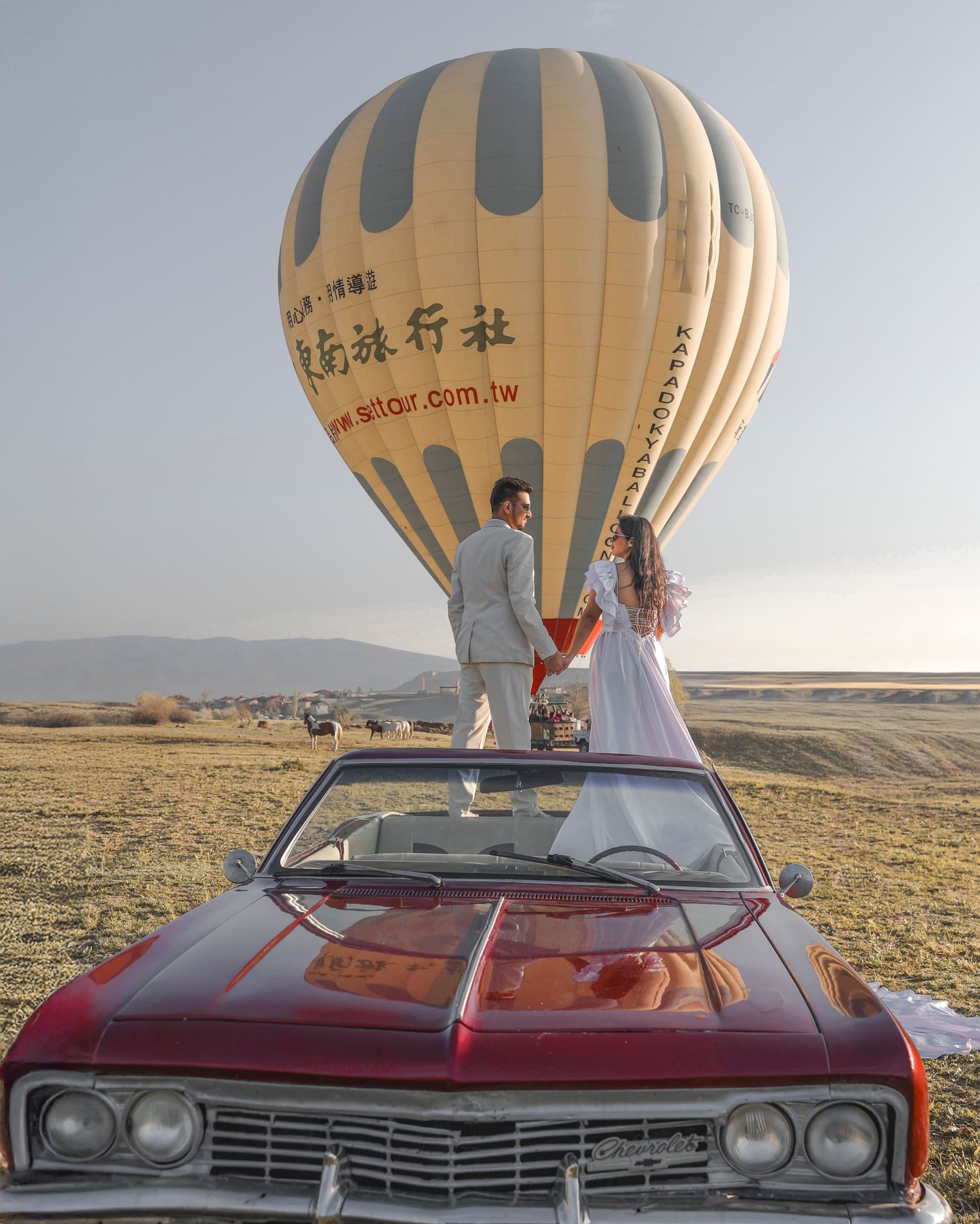 Couple in wedding attire standing on vintage red convertible car with hot air balloon in desert landscape.