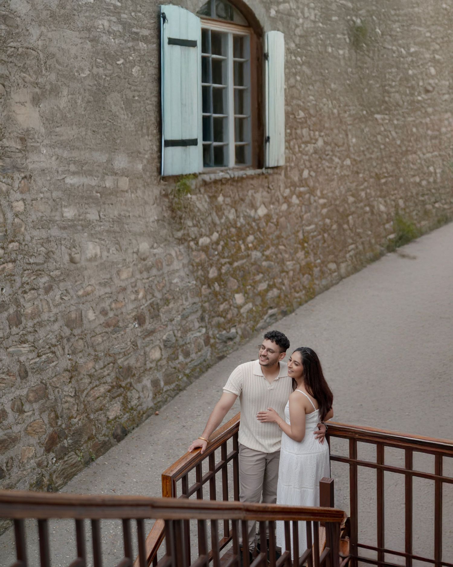 Couple embracing on stone staircase beside rustic wall with teal shuttered window in European setting.