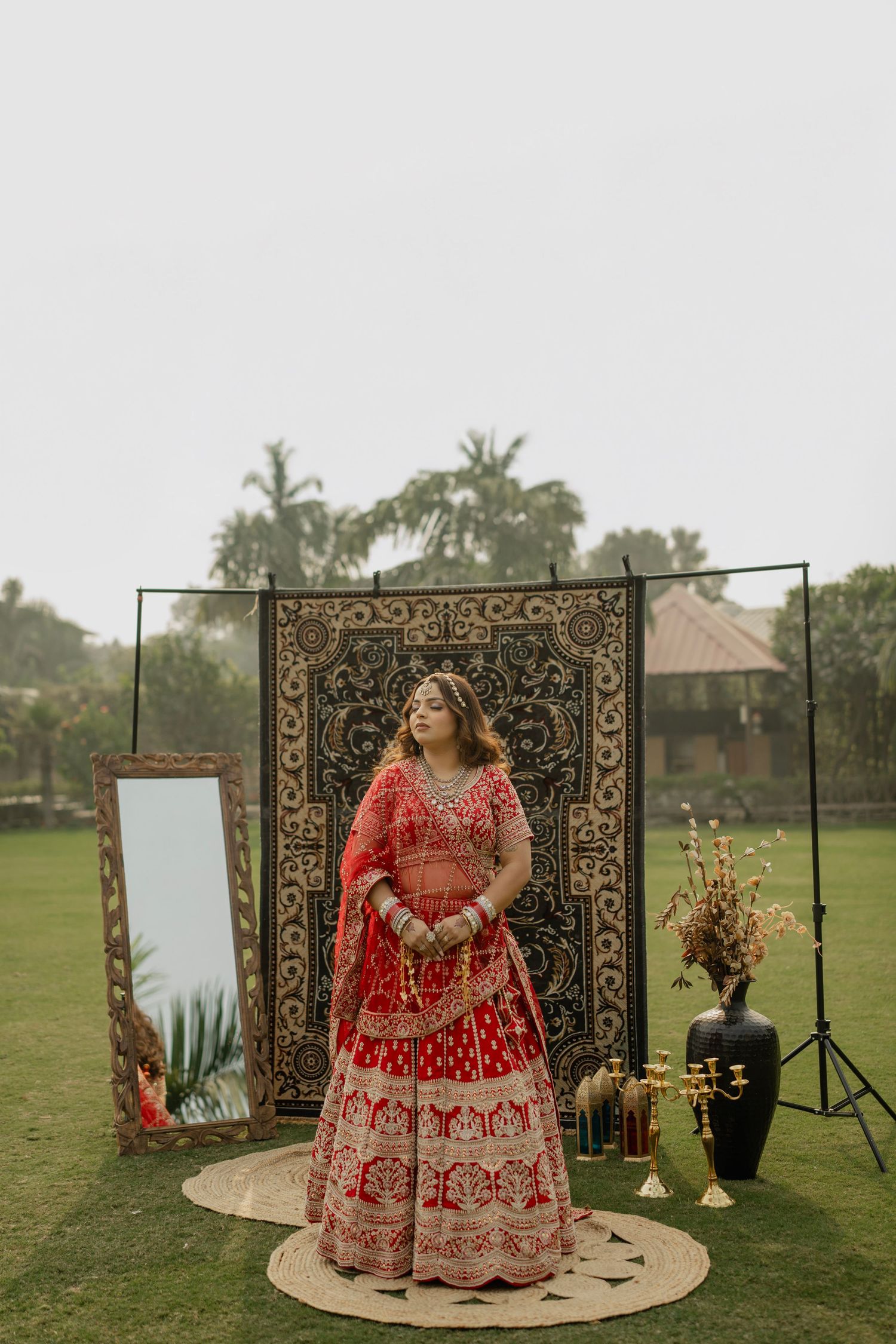 Bride in red embroidered lehenga posing outdoors at a garden wedding setup with decorative tapestry backdrop.
