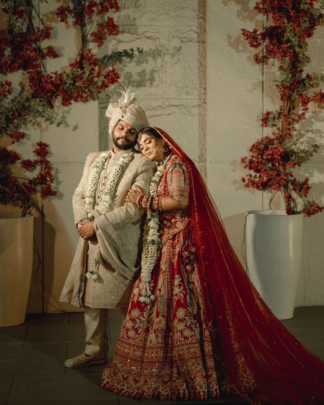 Indian bride in red lehenga and groom in cream sherwani pose together at traditional Hindu wedding ceremony.