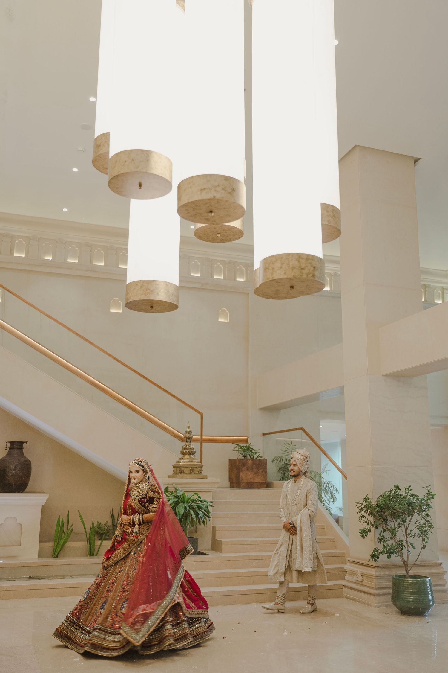 Indian bride in red lehenga and groom in cream sherwani stand in elegant white marble wedding venue lobby.