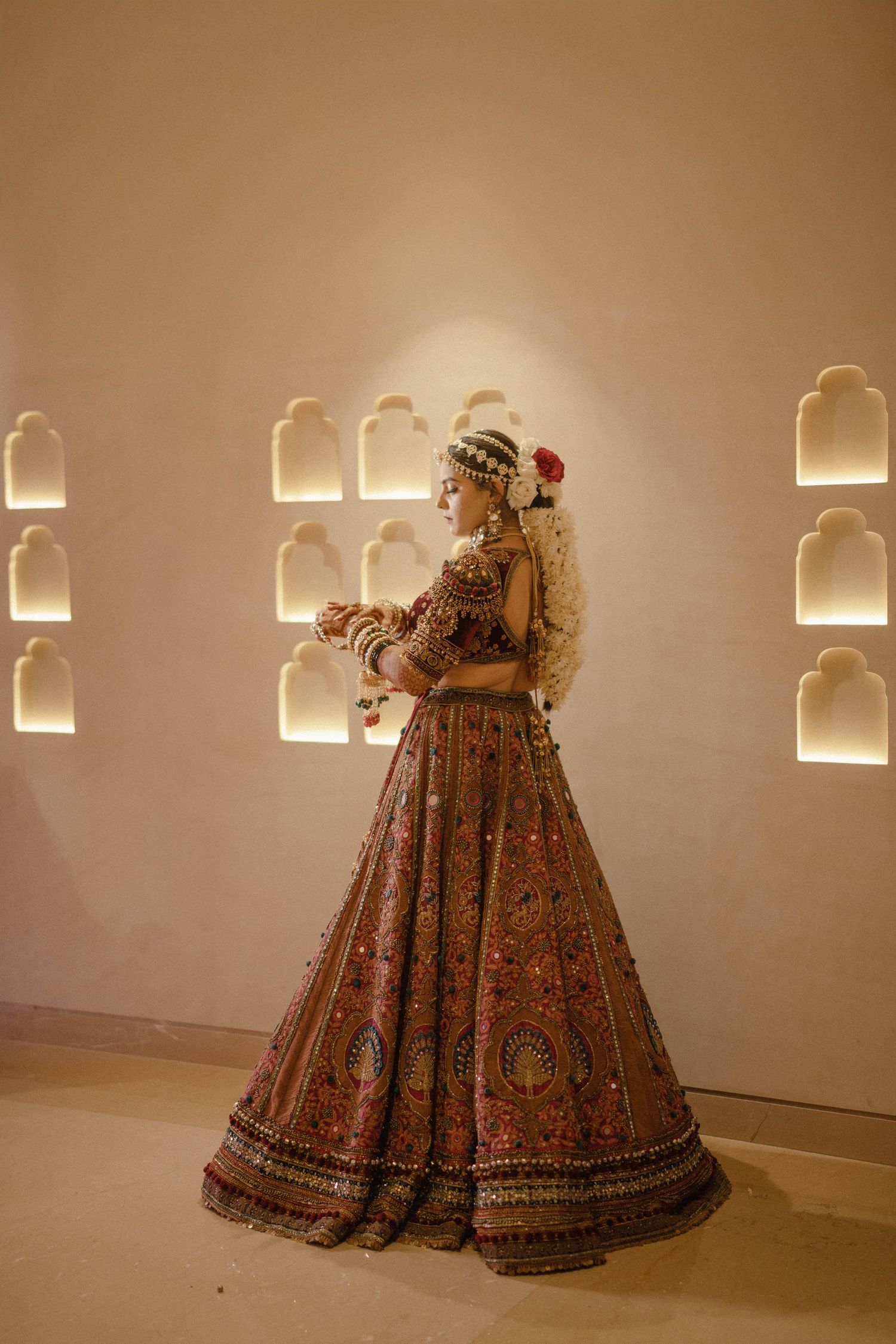 Bride in ornate brown embroidered lehenga with floral hair accessory poses elegantly in warmly lit arched alcove setting.