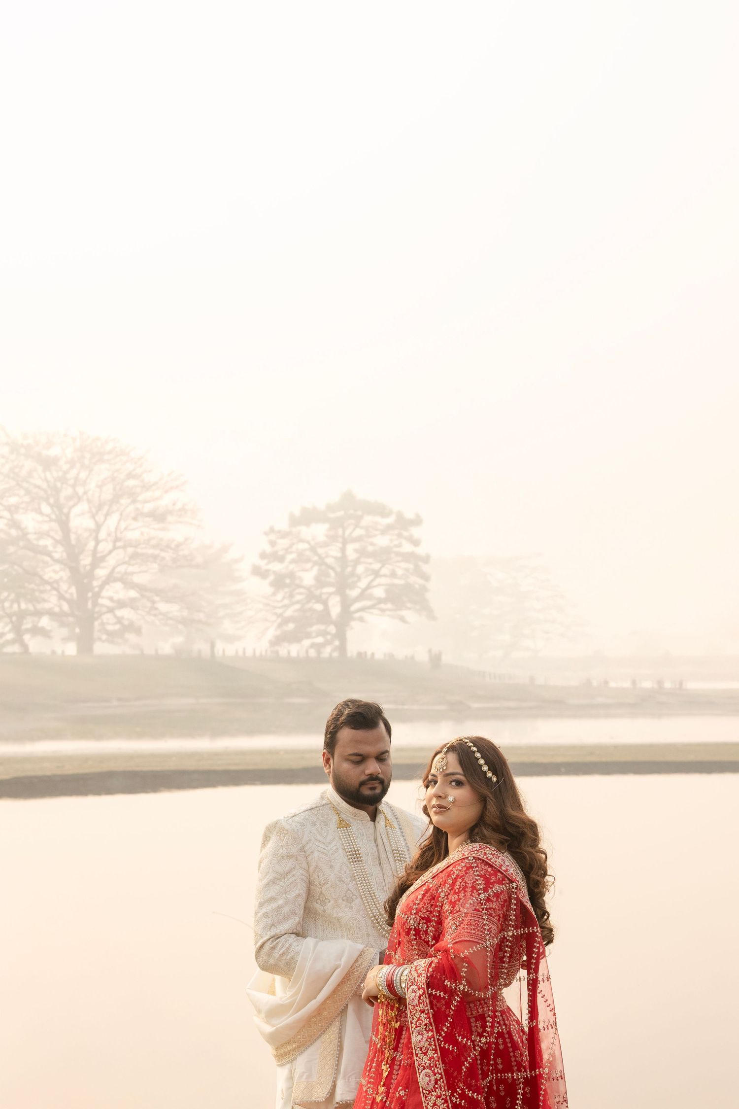 Indian couple in traditional wedding attire pose elegantly in a misty outdoor setting, bride in red lehenga, groom in ivory sherwani.