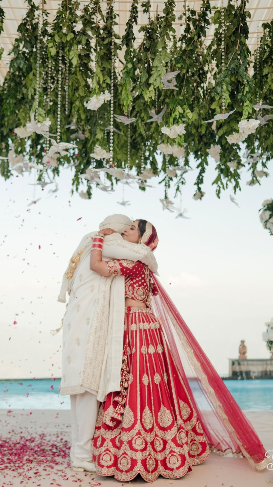 Indian bride in red lehenga and groom in white sherwani embrace under floral canopy with rose petals at wedding.