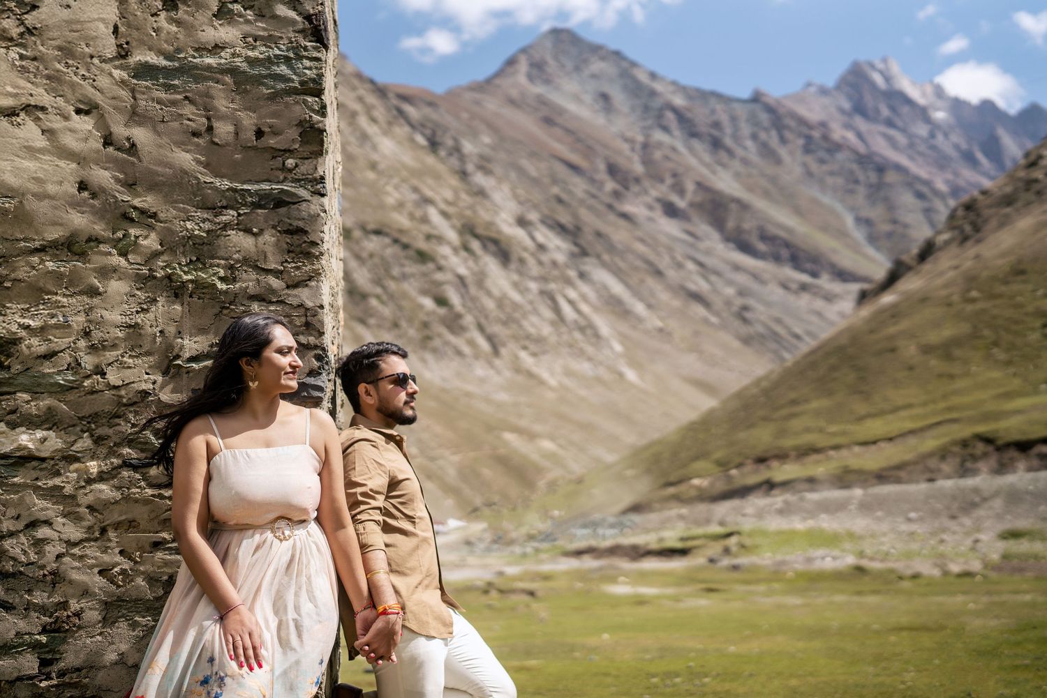 Couple in wedding attire holding hands against stone wall with scenic mountain valley backdrop.