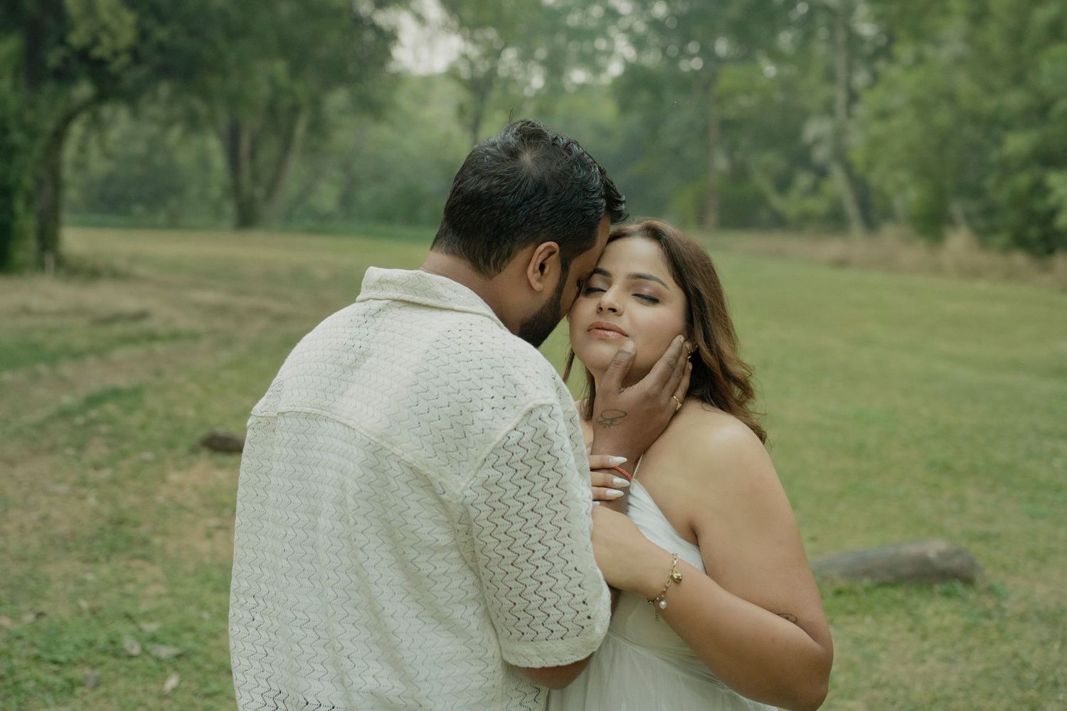 A couple shares a tender romantic moment in a lush green park, the man gently cradling the womans face.