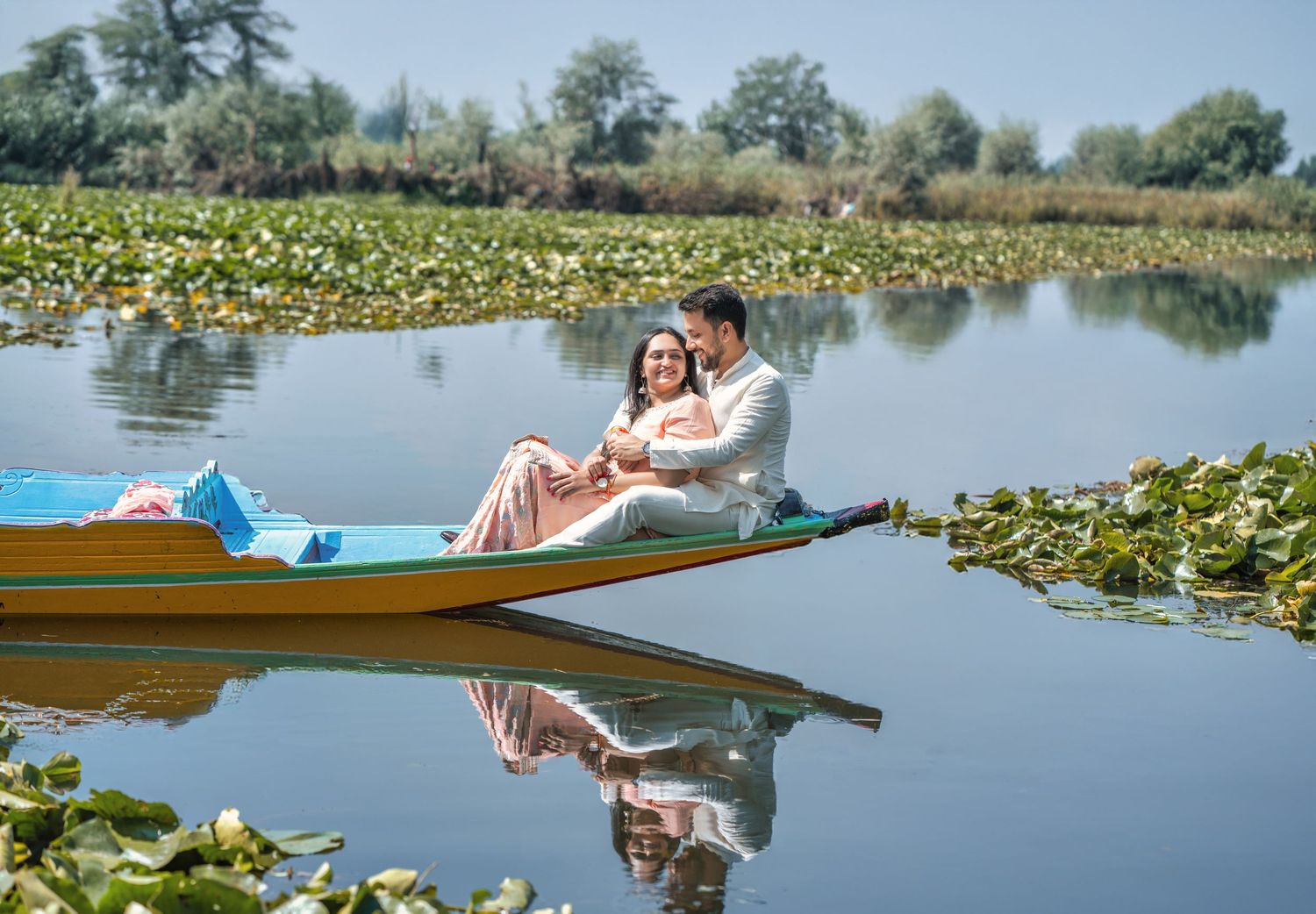 Couple sitting together in a colorful boat on a serene lake surrounded by lily pads, reflecting in calm water.