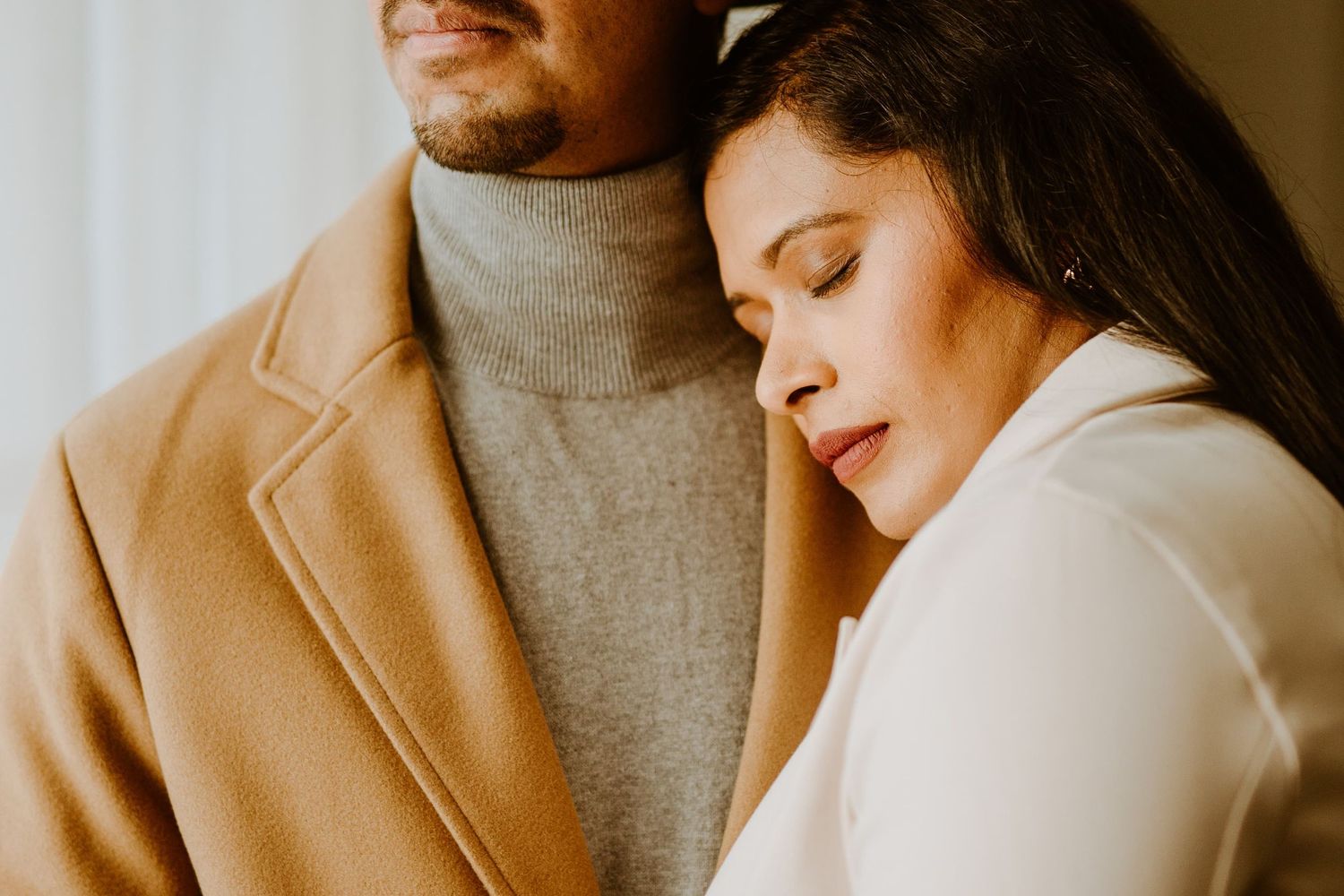 A woman rests her head on a man's chest, both dressed in elegant neutral tones, sharing a tender romantic moment.