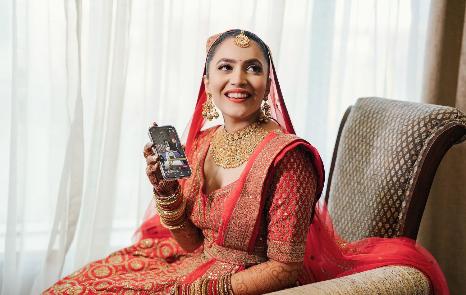 Bride in traditional red Indian attire seated on elegant gray chair with ornate jewelry.