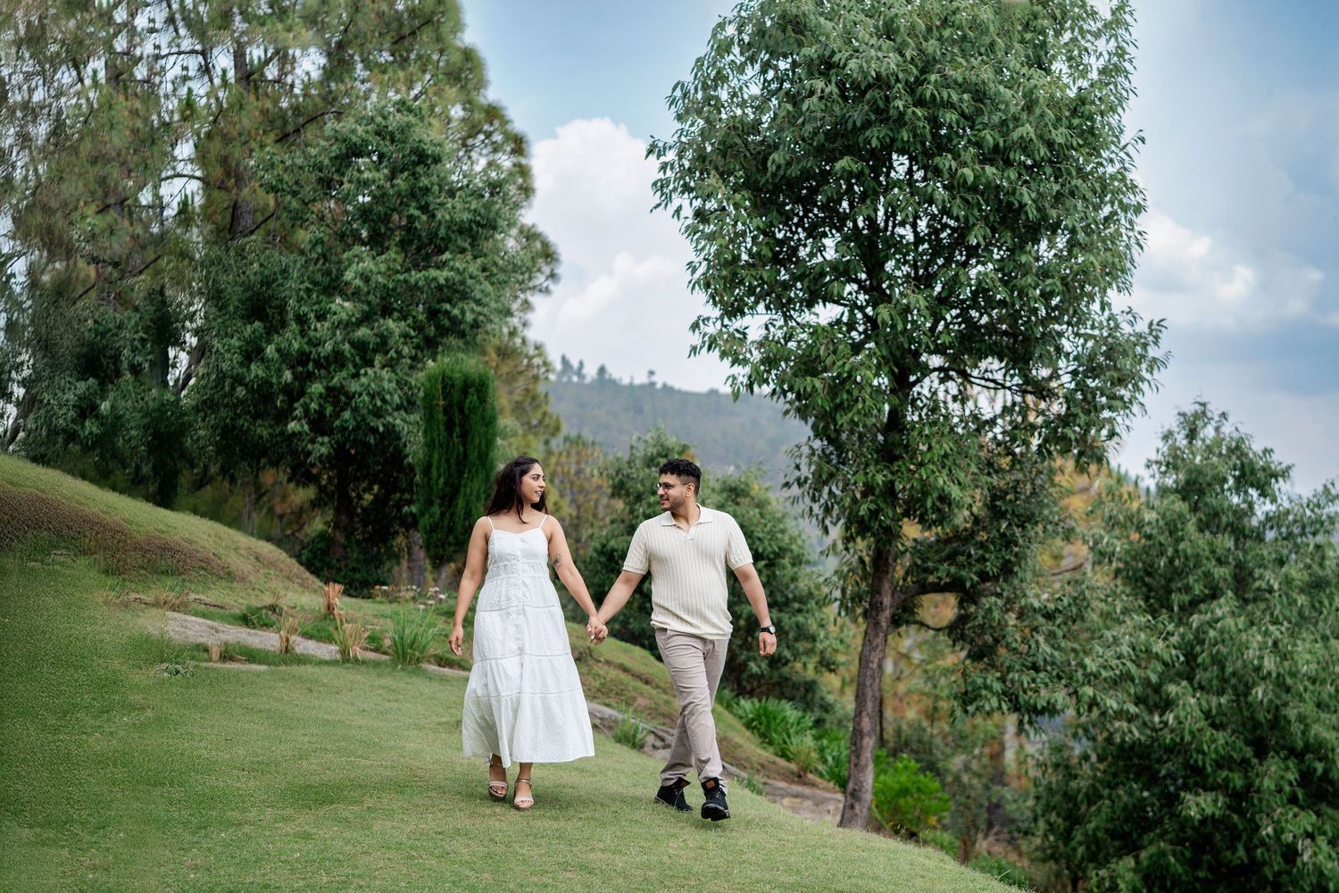 Couple walking through a lush green garden path with trees and grass on a sunny day.