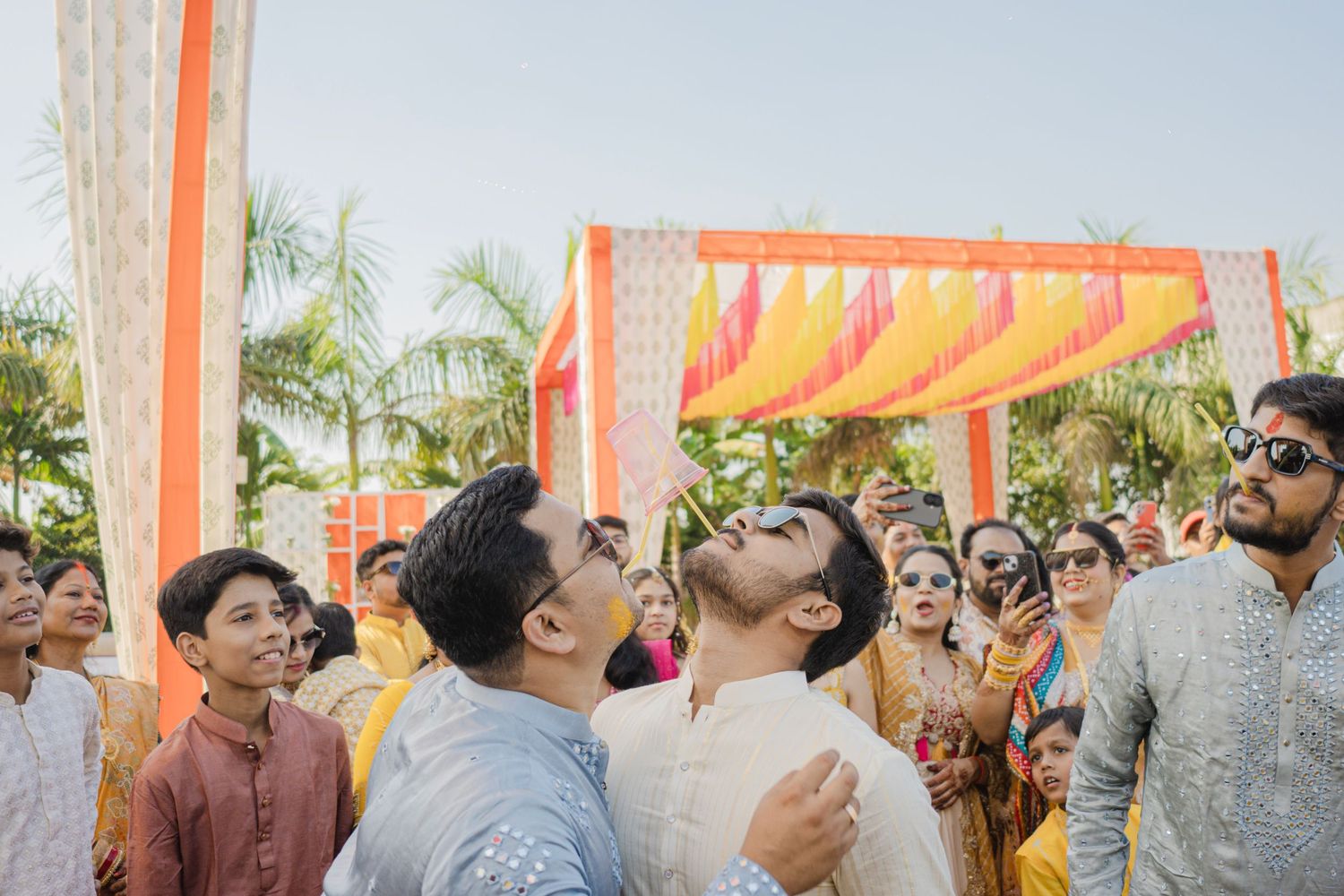 Two grooms share a joyful moment at a vibrant Indian wedding ceremony with colorful mandap decorations.