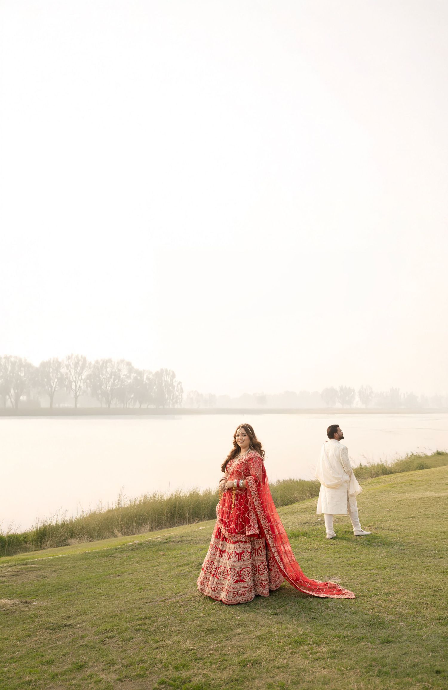 Bride in red lehenga stands by misty lakeside as groom in white kurta walks ahead during Indian wedding photoshoot.
