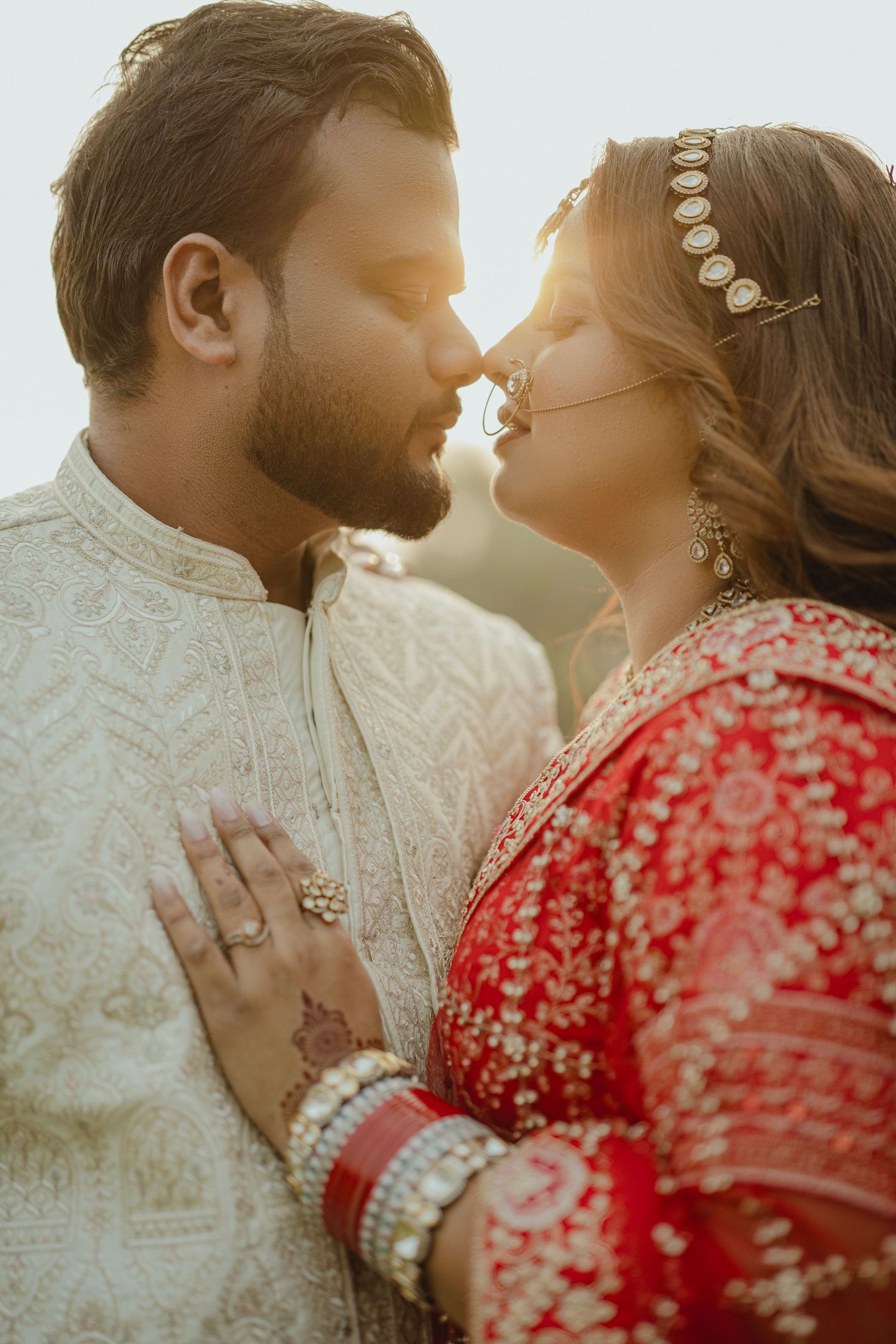 Indian bride in red lehenga and groom in white sherwani share a romantic moment during golden hour wedding shoot.