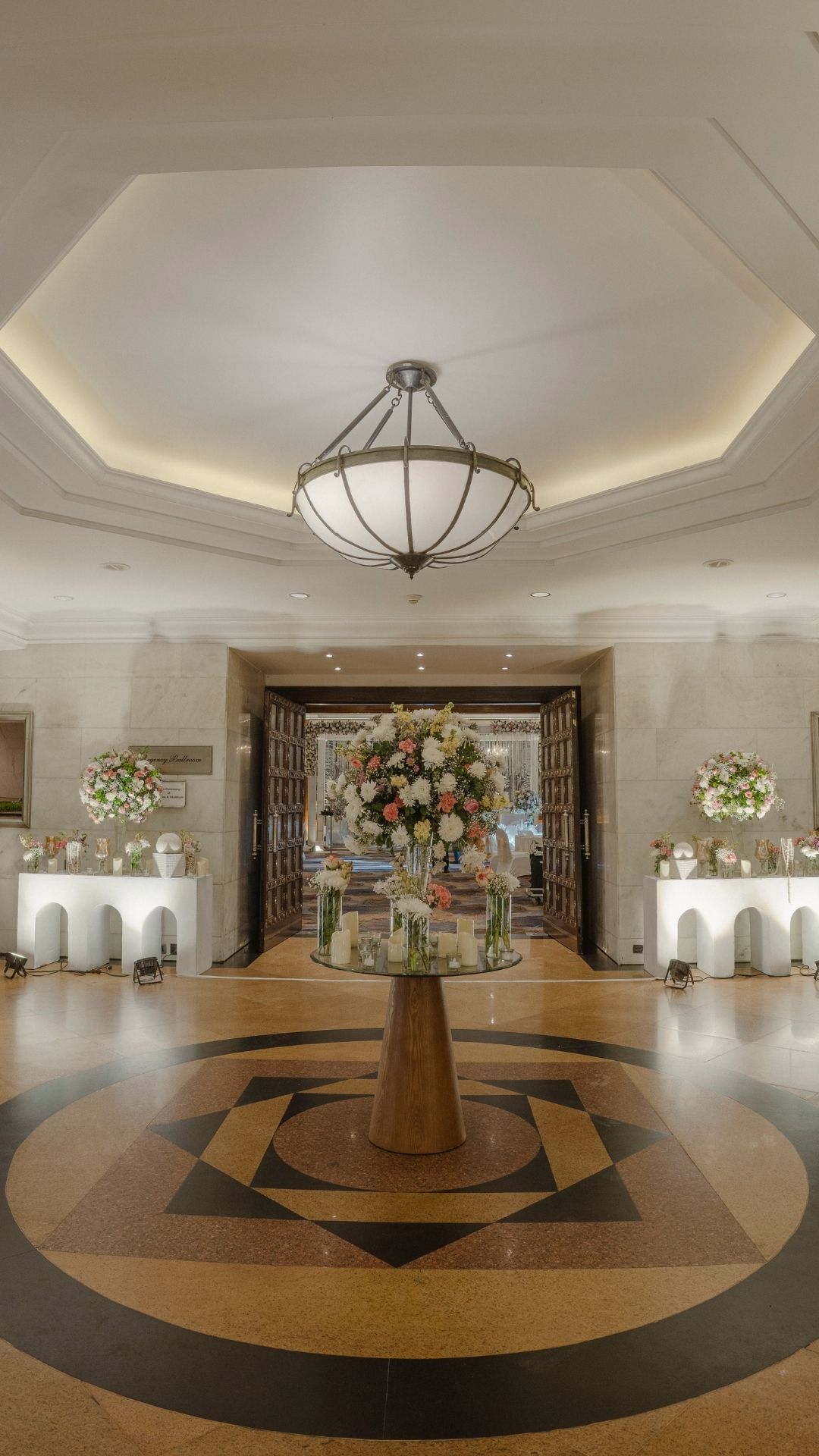 Elegant hotel lobby with marble floors, geometric ceiling, chandelier, and floral centerpiece on a round glass table.