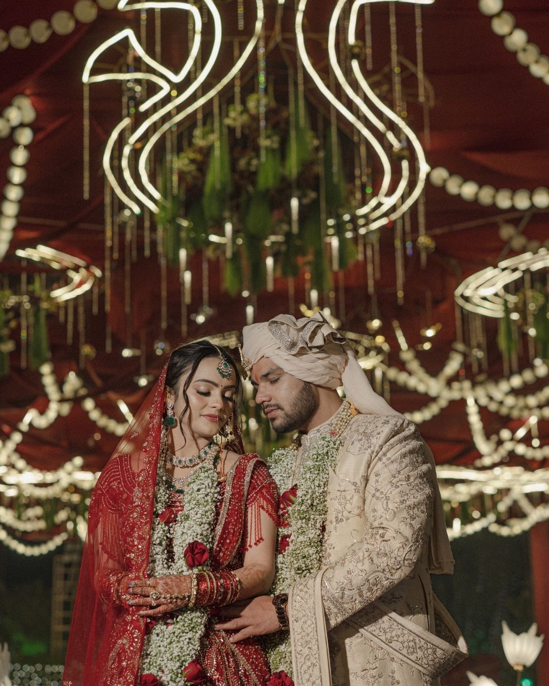 Indian bride in red lehenga and groom in ivory sherwani share an intimate moment under floral chandelier decorations.