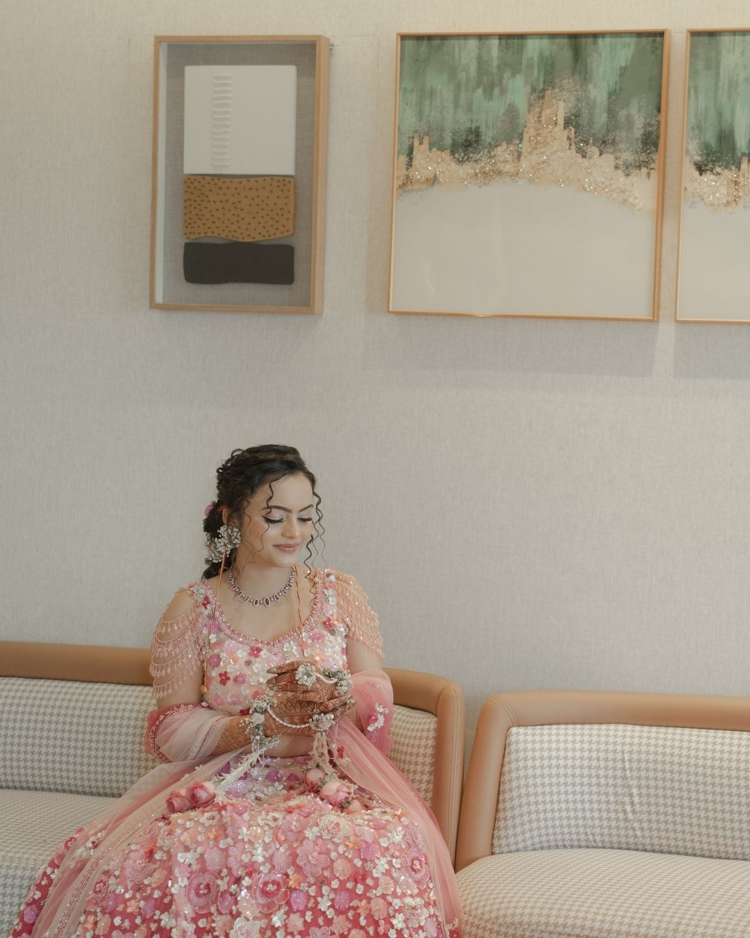Bride in pink floral lehenga sitting on sofa, wearing jewelry, with framed artwork on wall behind her.