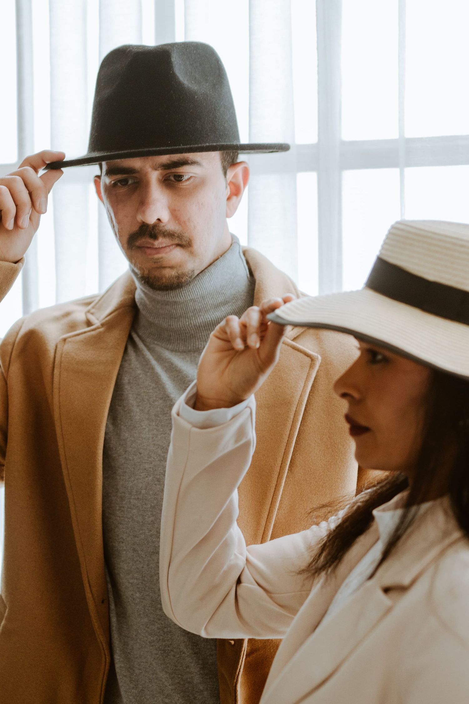 A stylish couple in beige coats and hats pose elegantly, with the woman adjusting the man's dark fedora.