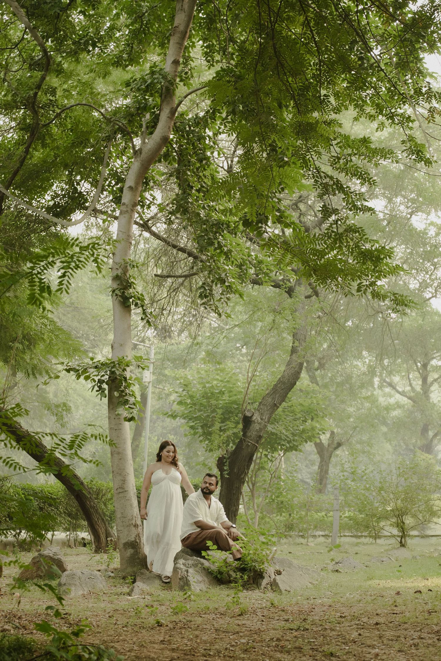 Couple in white outfits posing under lush green trees in a misty forest setting.