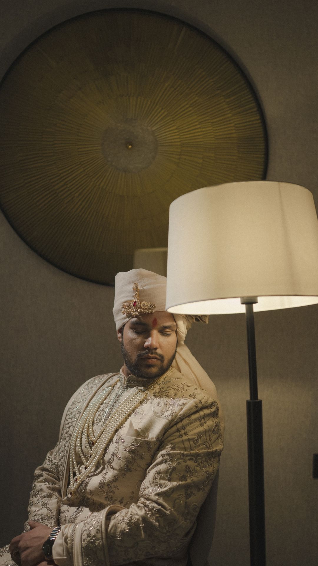 Indian groom in traditional white sherwani and safa turban seated beside a floor lamp with decorative wall art.
