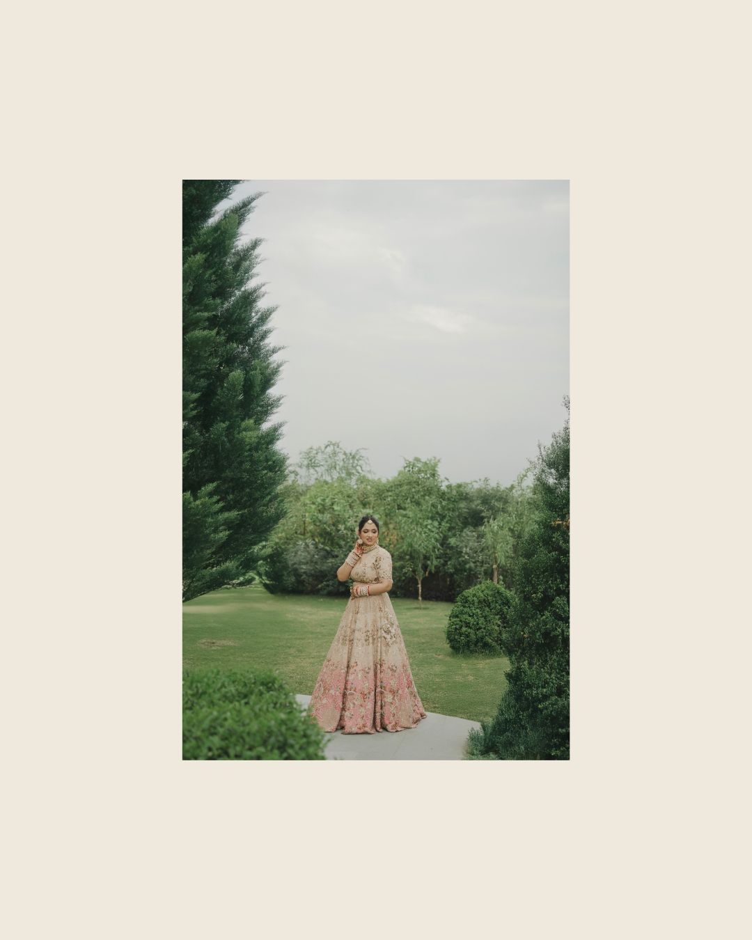 Bride in pink lehenga standing in lush green garden on overcast day.