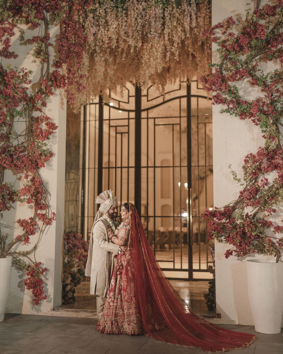 Indian bride in red lehenga and groom in cream sherwani pose under floral arch with pink bougainvillea blooms.
