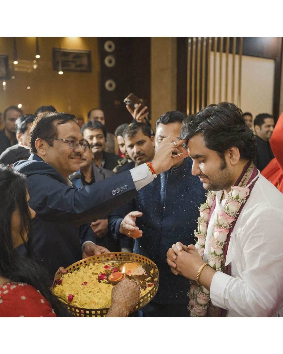 A groom receives tilak during a traditional Indian wedding ceremony surrounded by guests.