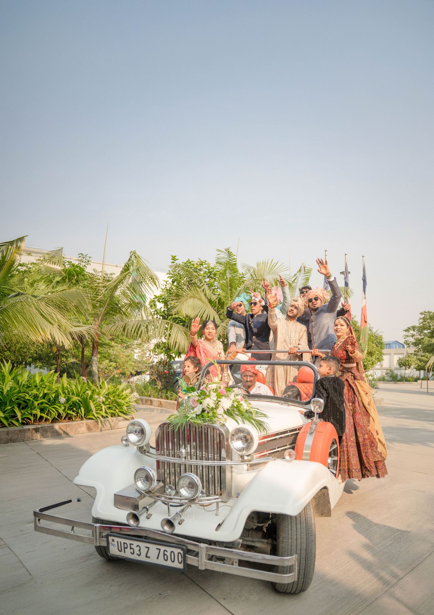 Wedding party celebrating in a vintage white classic car decorated with flowers, raising hands joyfully outdoors.