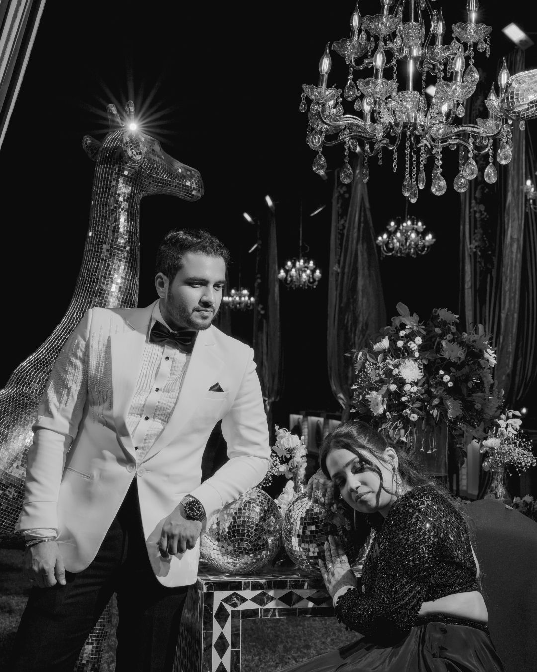 Black and white photo of elegant couple in formal wear posing with disco ball under crystal chandelier.