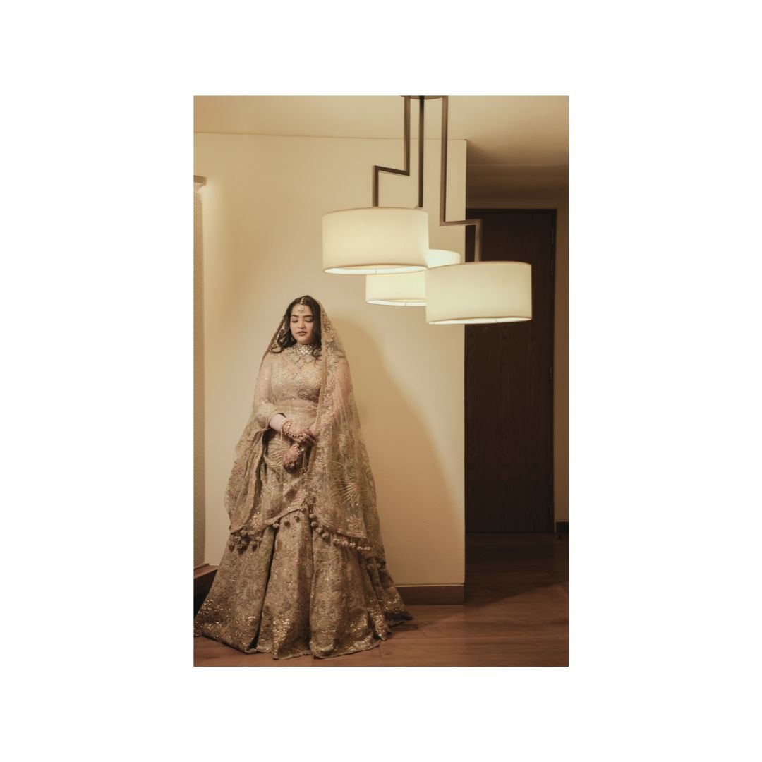 A bride in an ornate golden lehenga stands elegantly in a warmly lit room with modern pendant lights.